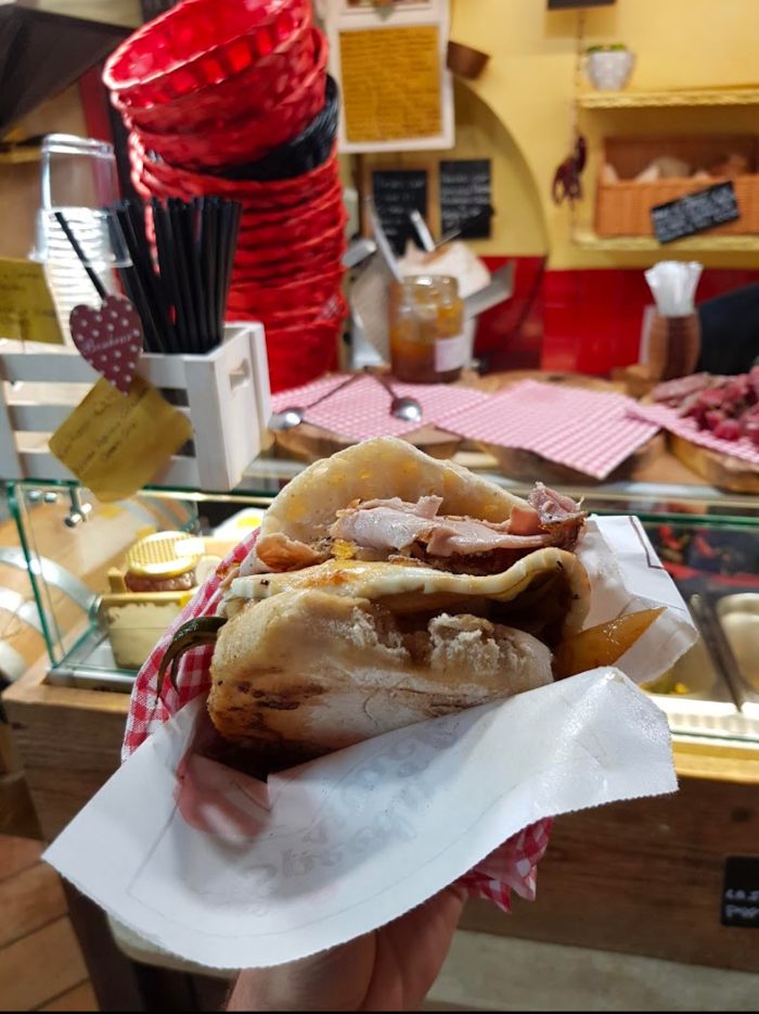 Panino farcito con salumi e altri ingredienti, avvolto in un tovagliolo e tenuto in mano, in una bottega con decorazioni rustiche sullo sfondo